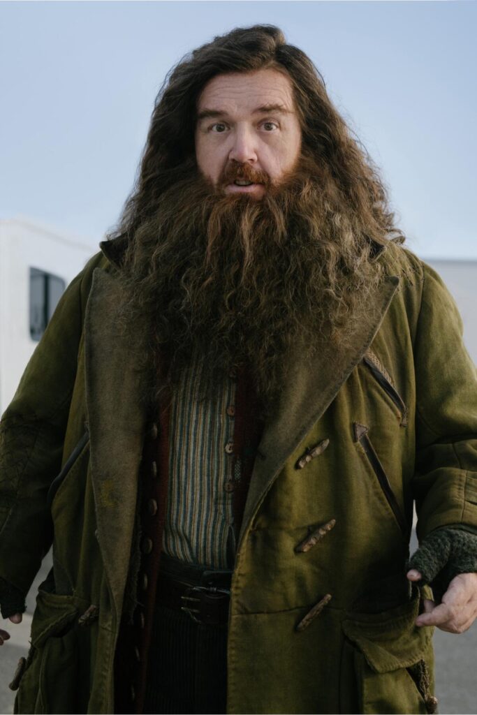 A man with long hair and a long shaggy beard clad in an olive drab coat over a striped shirt and dark pants