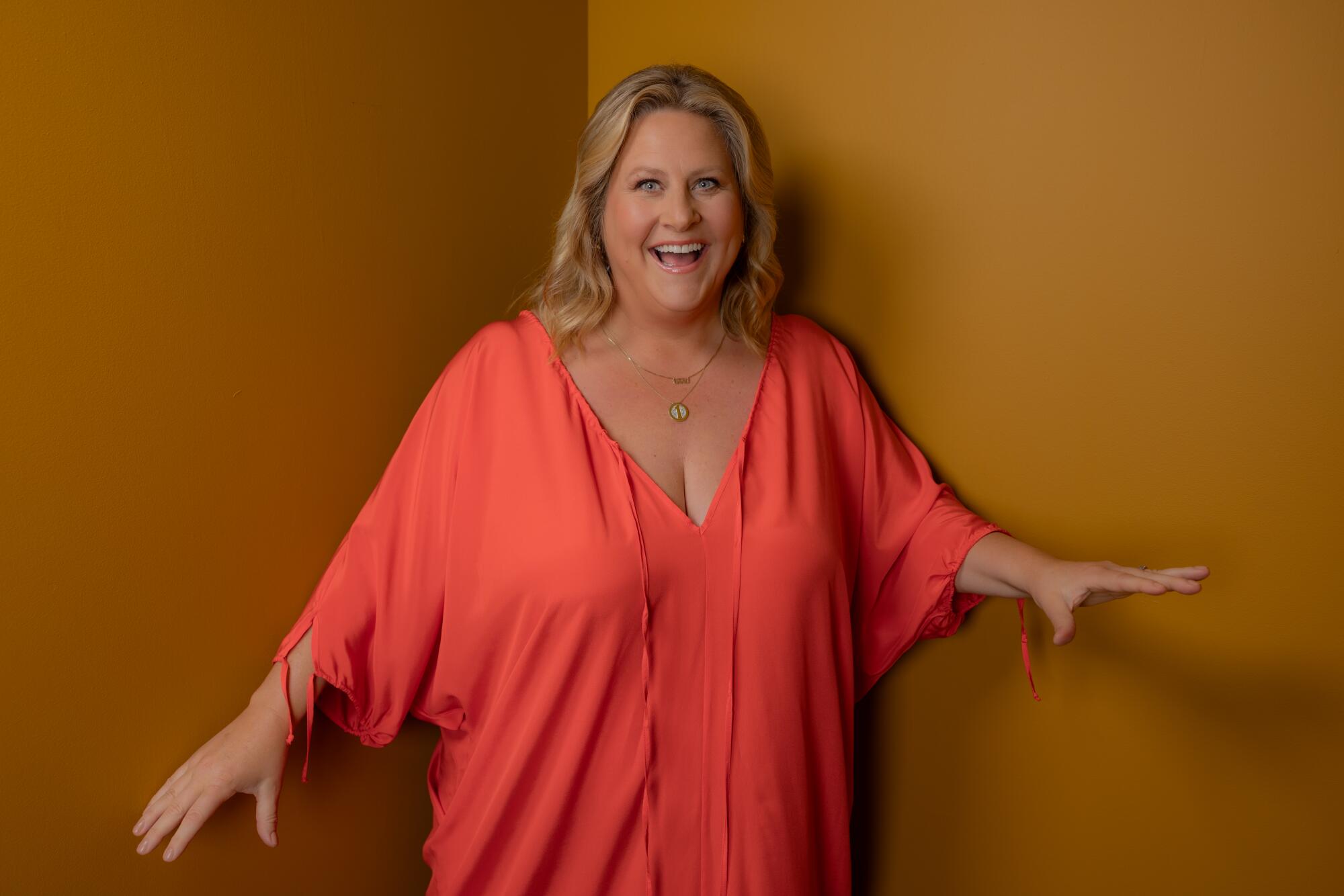 Bridget Everett poses with a smile and her arms out to her sides against a wal.