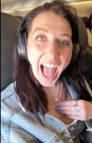 Woman on airplane excitedly showing necklace after receiving compensation for flight overbooking.