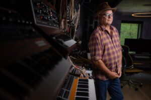 Man in a fadora and glasses leaning against a synthesizer