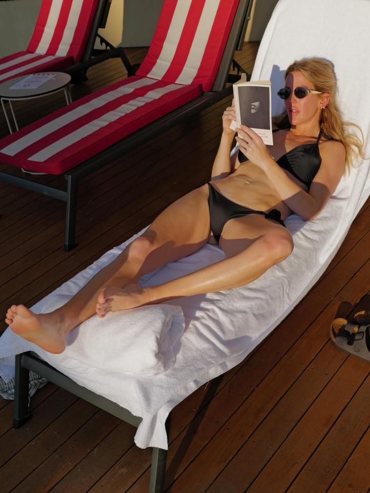 Woman in a bikini reading a book on a lounge chair.