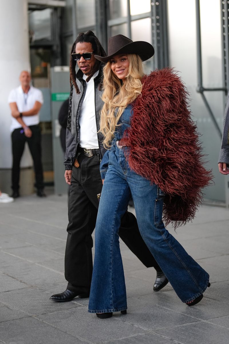 Jay-Z wears a black leather jacket with a Louis Vuitton monogram logo print, a white T-shirt, a belt, black wide-leg pants, and black leather pointed shoes.