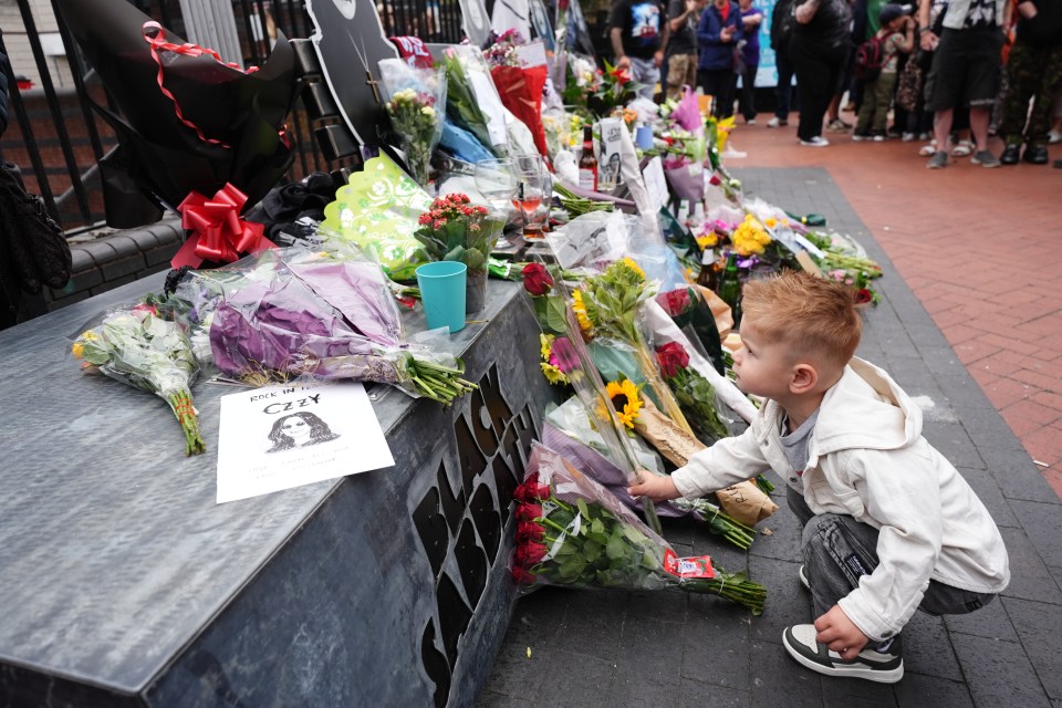 Criança colocando uma rosa entre as flores deixadas em um memorial do Black Sabbath.
