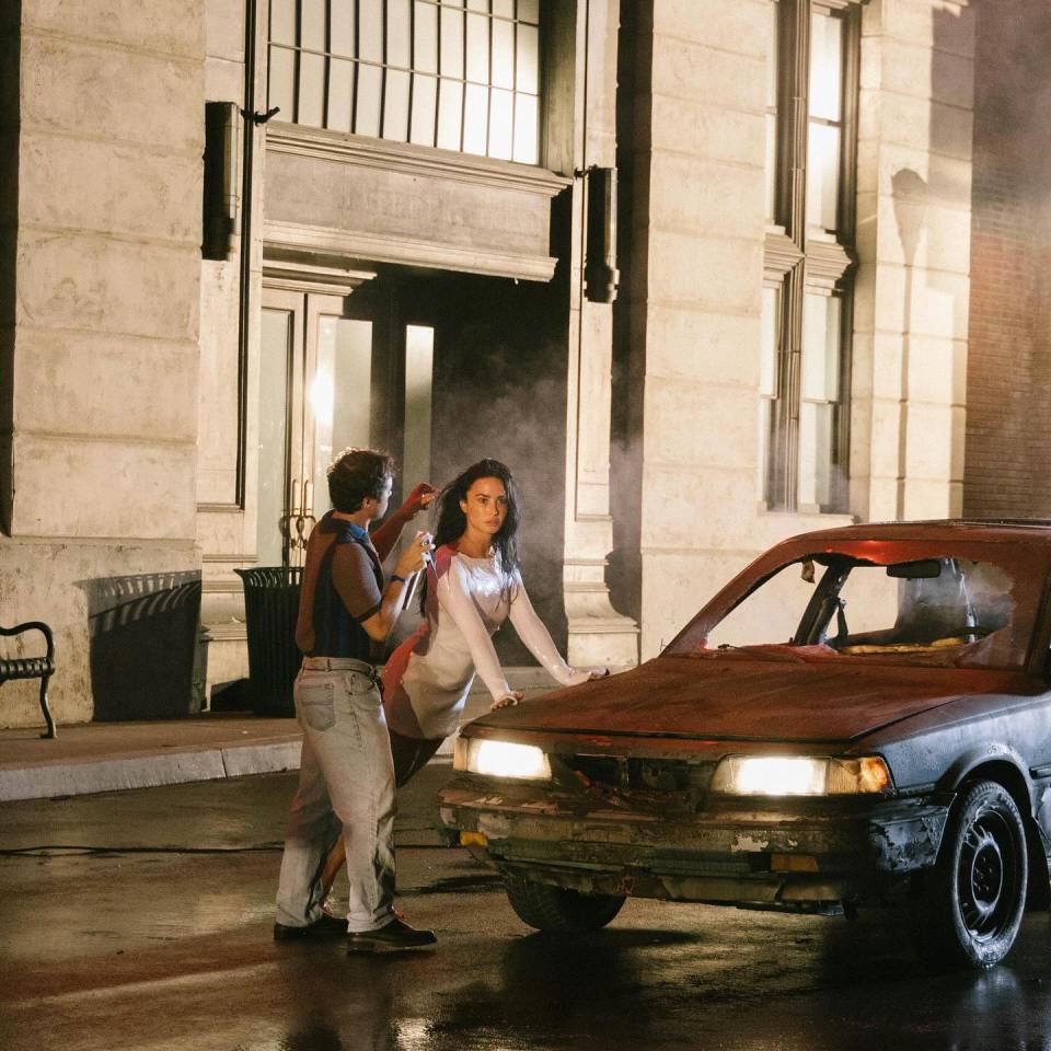 Demi Lovato behind a damaged car with a crew member.