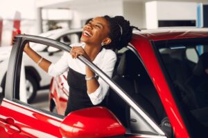 A happy young woman getting into a new red car in a car dealership.