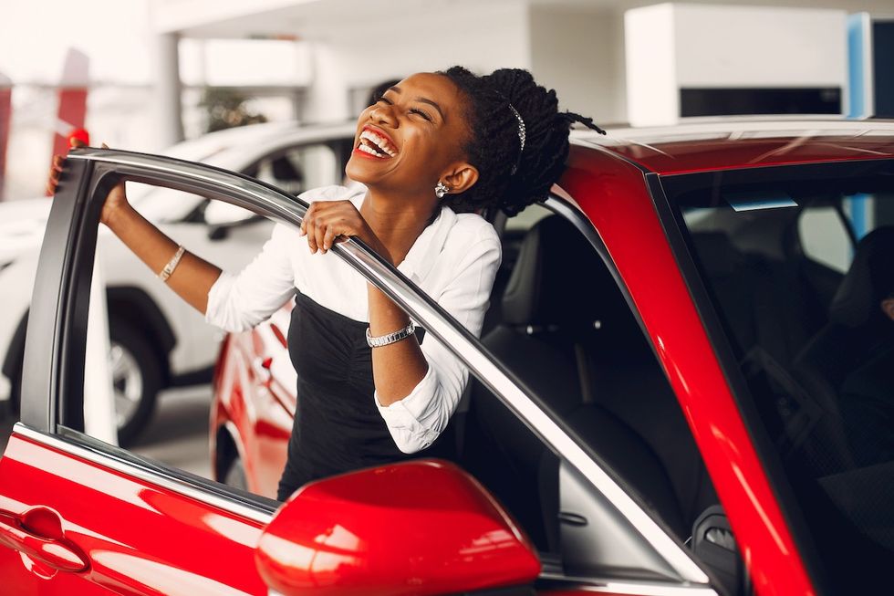 A happy young woman getting into a new red car in a car dealership.