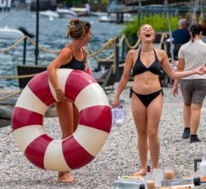 Two women in bikinis laughing on a beach; one carries a large inflatable ring.