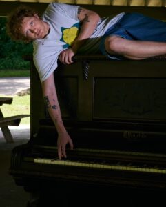 In a covered picnic area of Shelby Park, Knox lays on an upright piano, with his right arm extended down to play a key.