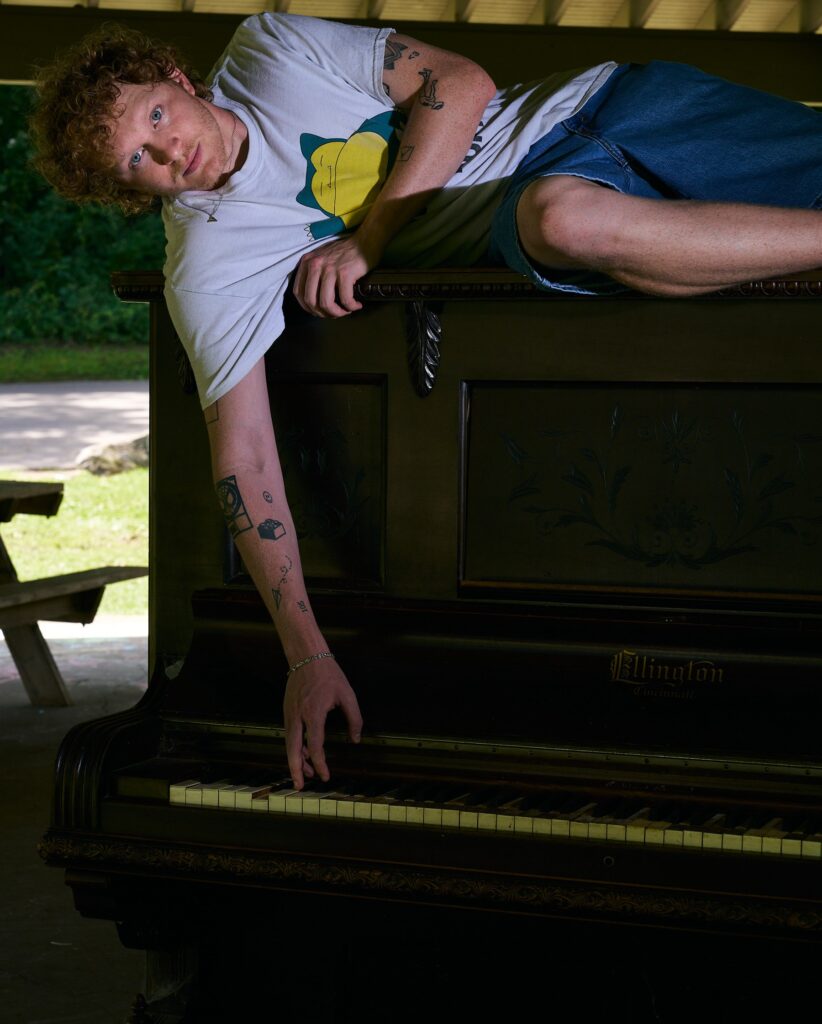 In a covered picnic area of Shelby Park, Knox lays on an upright piano, with his right arm extended down to play a key.