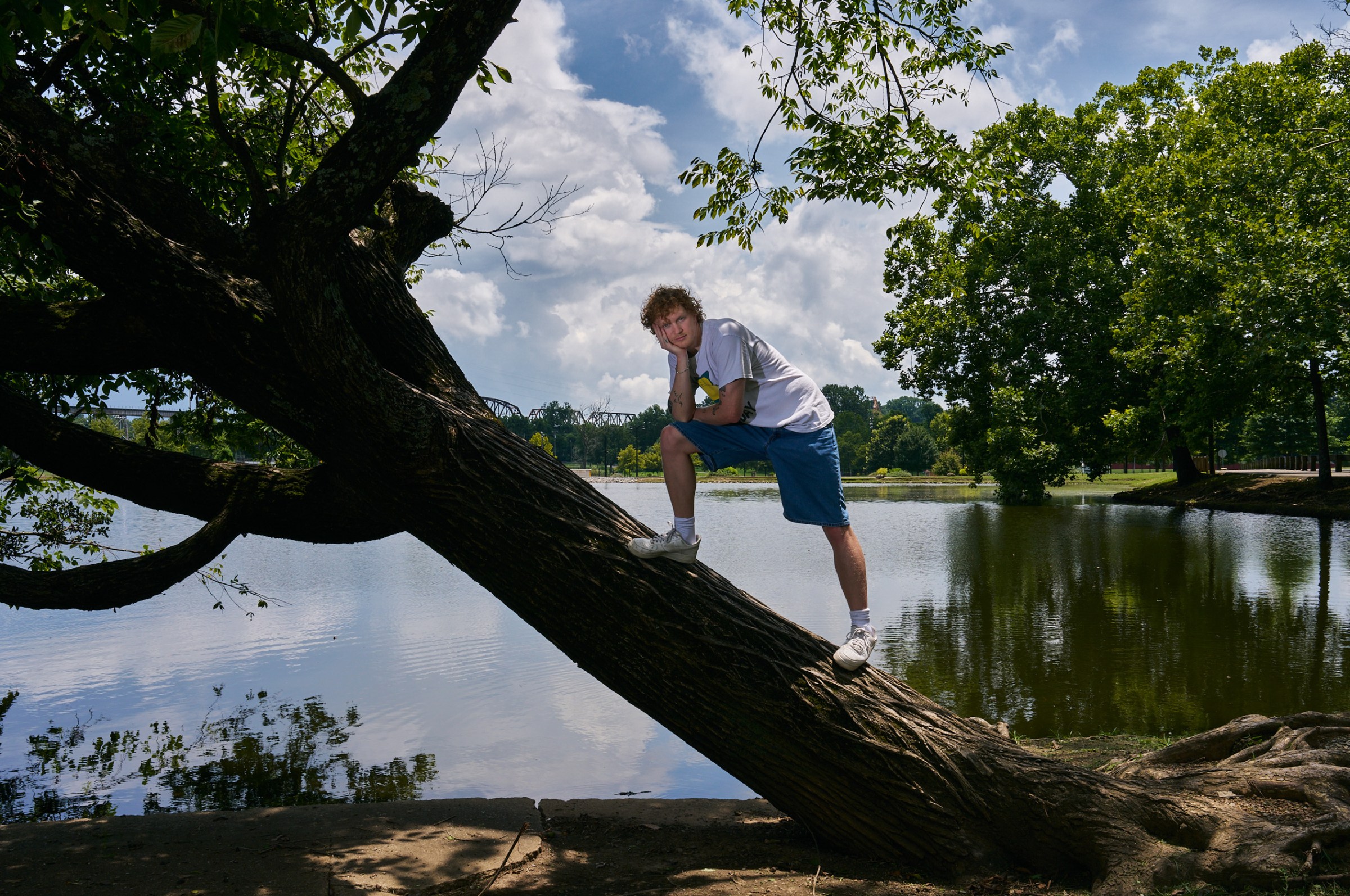 On a bright sunny day Knox Morris in a loose white t-shirt and jean shorts holds his head in his hand while leaning on a knee atop a large tree growing almost parallel to the ground. Behind him is a body of water at the edge of a lake.