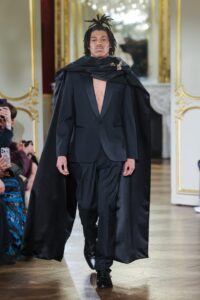 Model in black tuxedo and cape walking a runway.