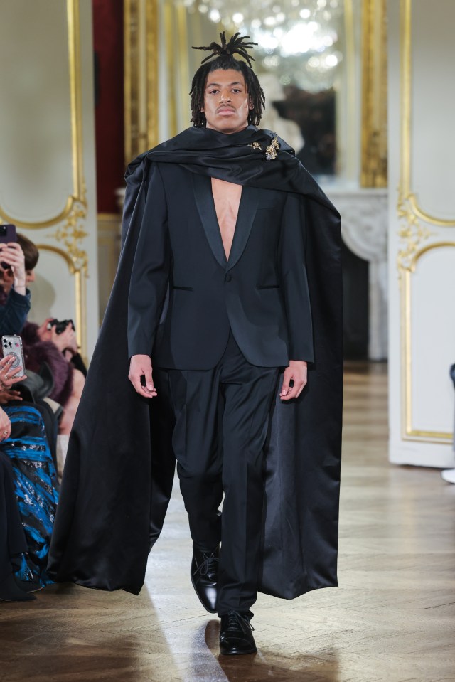 Model in black tuxedo and cape walking a runway.