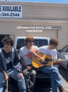 Three young men sitting on the back of a truck, playing a guitar; announcement for upcoming performance opening for Royel Otis.