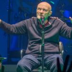 Phil Collins, seated and speaking into a microphone at a concert.