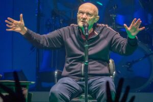 Phil Collins, seated and speaking into a microphone at a concert.