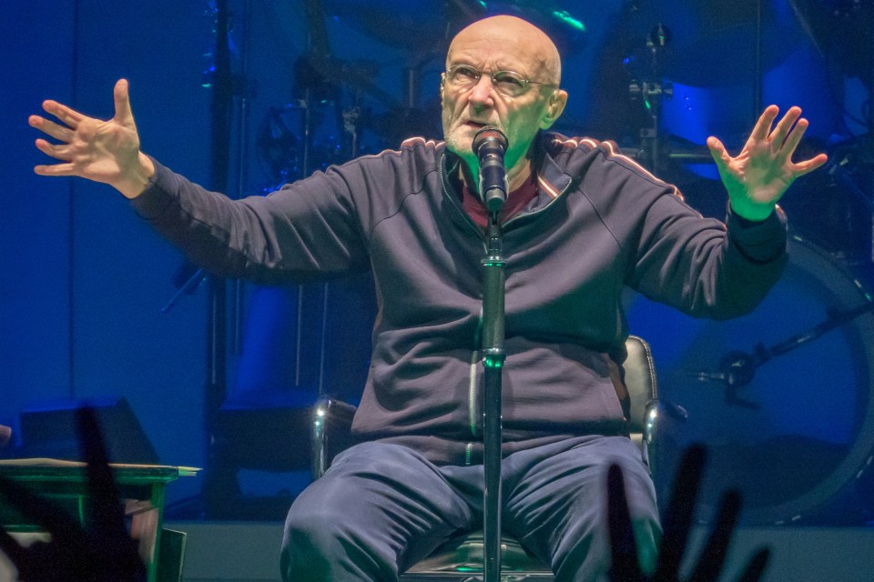 Phil Collins, seated and speaking into a microphone at a concert.