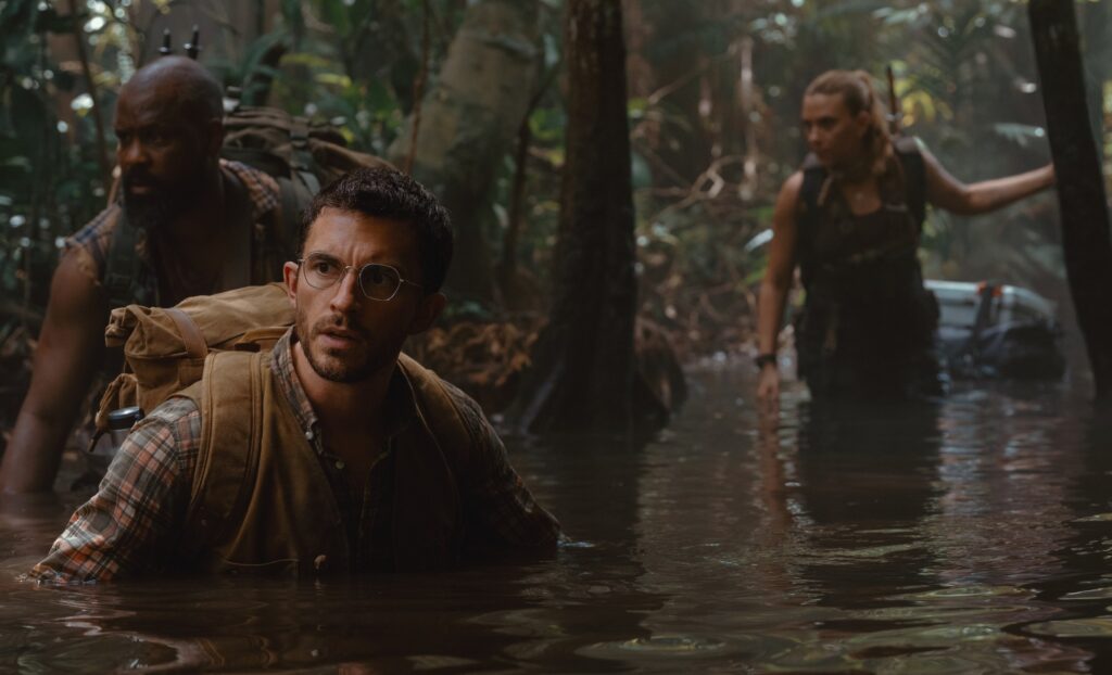 Two men (Bechir Sylvain and Jonathan Bailey) wade chest-deep through a swamp in Jurassic World Rebirth as Scarlett Johansson follows, only thigh deep in the water behind them
