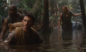 Two men (Bechir Sylvain and Jonathan Bailey) wade chest-deep through a swamp in Jurassic World Rebirth as Scarlett Johansson follows, only thigh deep in the water behind them