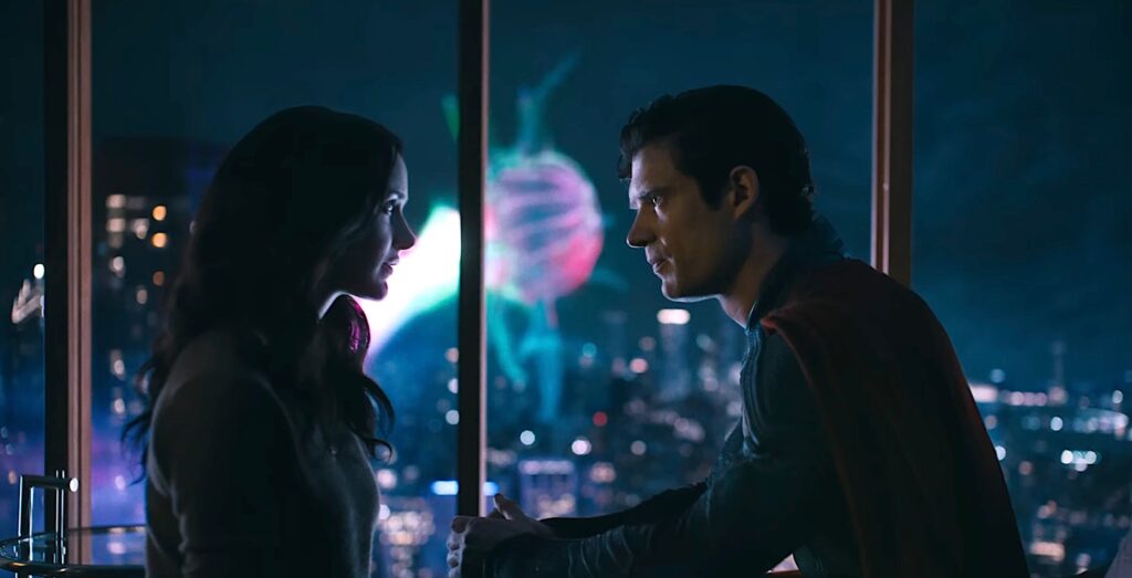 Superman (David Corenswet) and Lois Lane (Rachel Brosnahan) sit close together, across from each other, in a darkened high-rise apartment room, while a giant glowing flying jellyfish creature floats outside the window behind them in James Gunn’s Superman