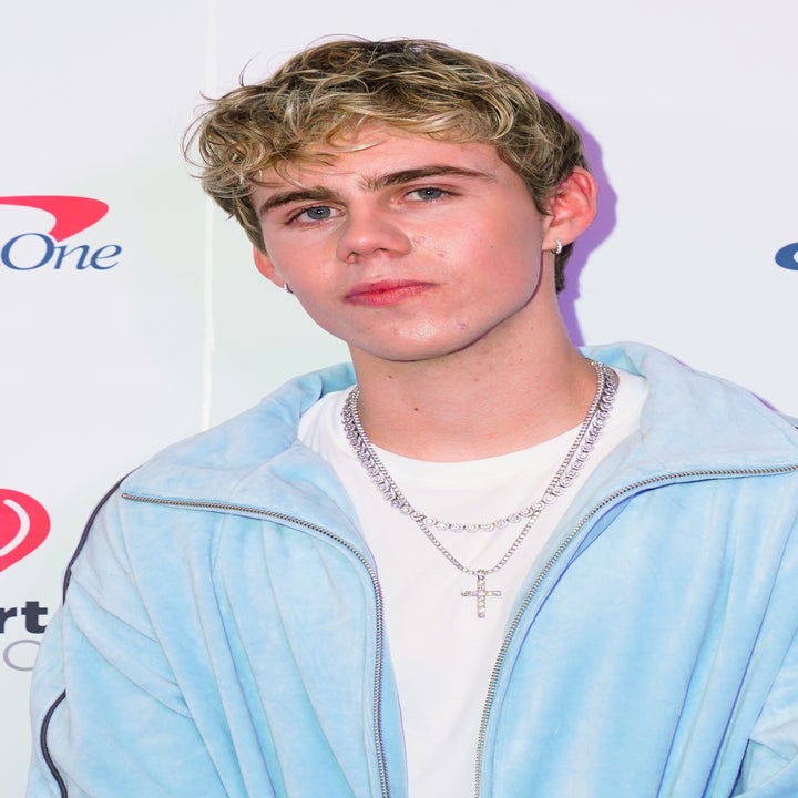 Young man in casual fashion wears a light jacket over a T-shirt with layered necklaces at an iHeartRadio event backdrop