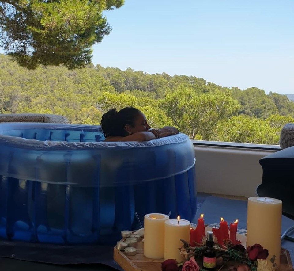 Person relaxing in a birthing pool overlooking a scenic landscape with candles and flowers nearby.