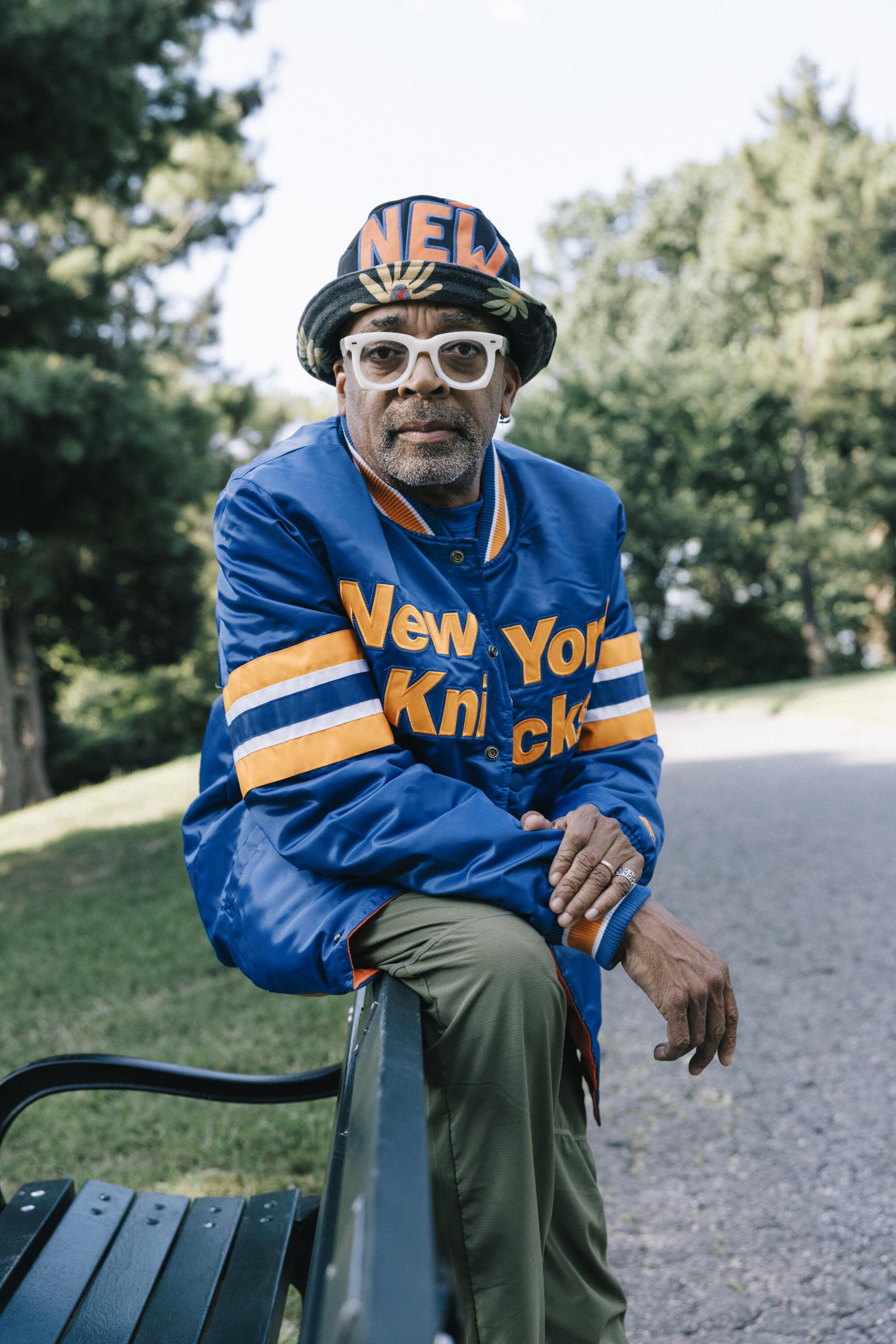 A man in shades and Knicks gear stands by a bench, looking into the lens.