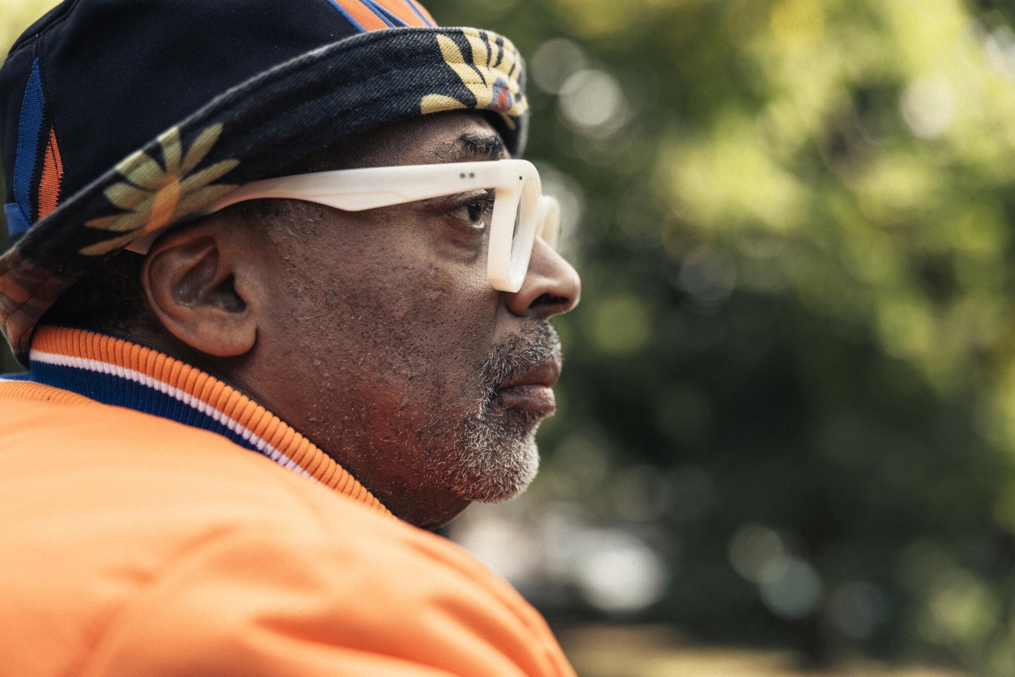 A man in Knicks gear and thick white-rimmed glasses looks to the side in profile.