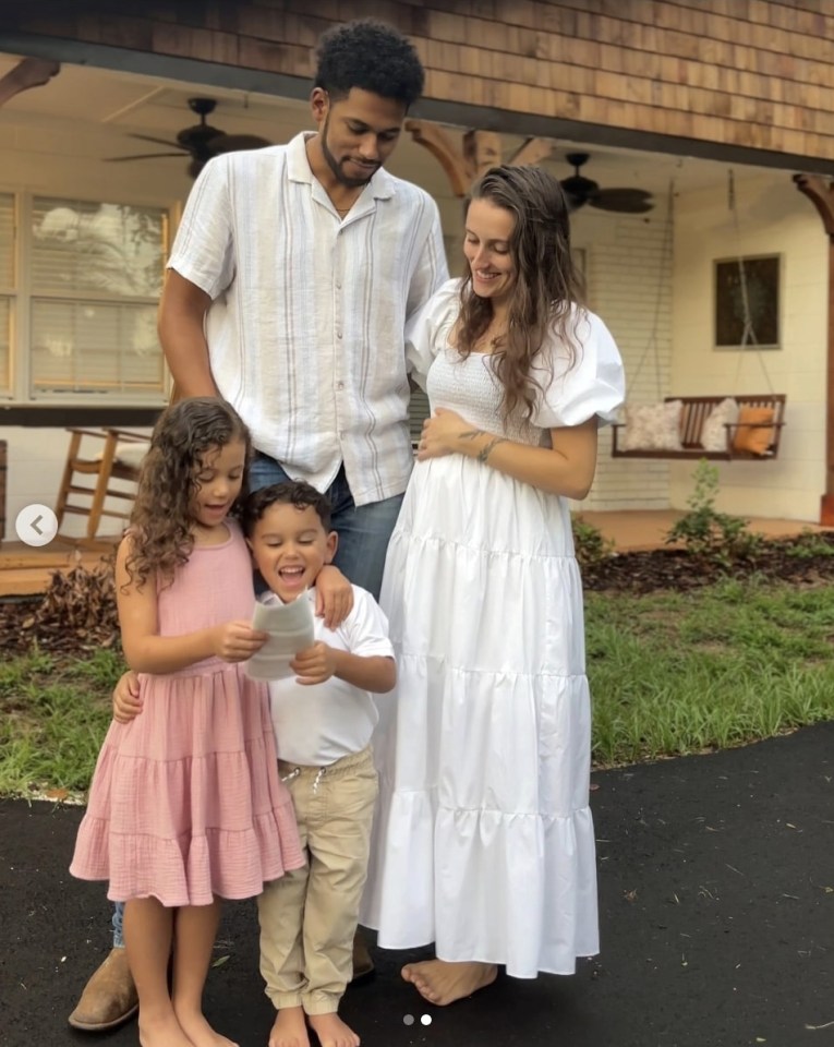 Family of four outside their home; the pregnant mother is holding her belly, the father is standing next to her, and their two children are holding a piece of paper.
