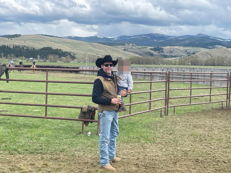 Man in cowboy hat holding a child, standing by a fence with cattle and a mountainous background.