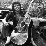 Black and white photo of Bobby Whitlock, songwriter and guitarist, posing with an acoustic guitar.
