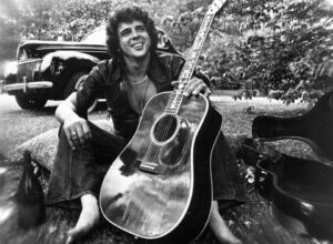 Black and white photo of Bobby Whitlock, songwriter and guitarist, posing with an acoustic guitar.