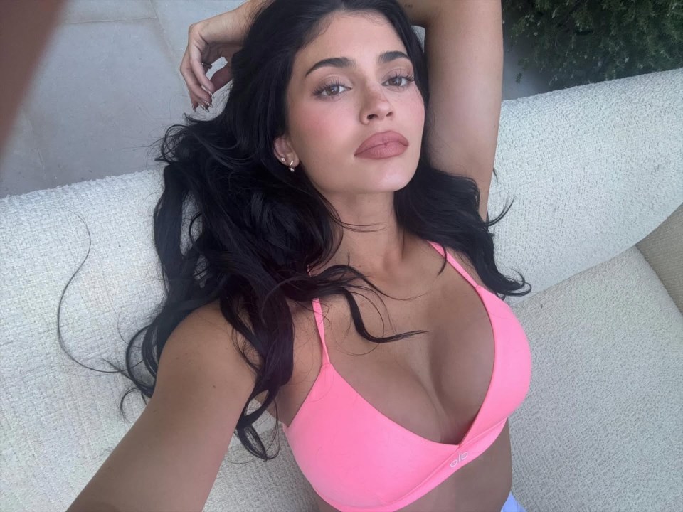 Kylie Jenner em um sutiã esportivo rosa, relaxando em um sofá.